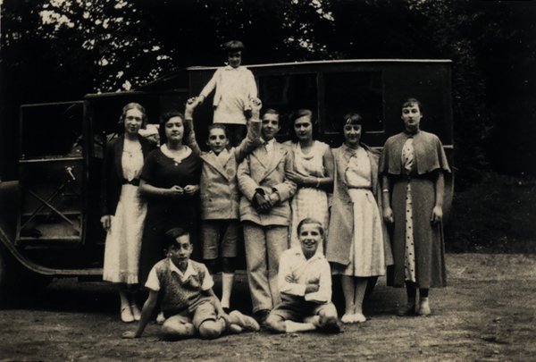 Groupe d'enfants et d'adultes posant devant un ancien véhicule. Noir et blanc.ᴬᴵ