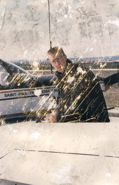 Homme blond à lunette devant une voiture. Image avec rayures.