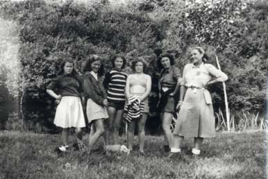 Six jeunes filles debout alignées dans l’herbe, arbres en arrière-plan.