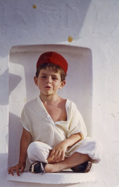 Petit garçon à toque rouge assis en tailleur dans la niche d’un mur blanc.