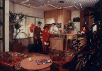 Homme et femme debout devant la réception d’un Hôtel, table et fauteuils en avant-plan.