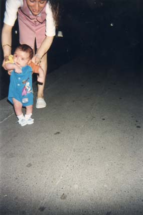 Petit enfant tenu debout en position de marche par une femme en arrière-plan.