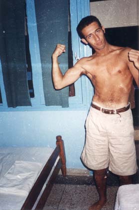 Homme brun torse nu en short montrant les muscles de ses bras dans une chambre.