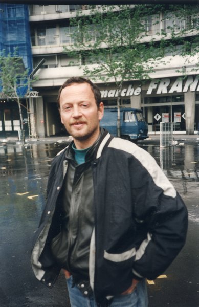 Homme à barbe claire en blouson, debout les mains dans ses poches de jeans, devanture d’un « Franprix » en arrière-plan.