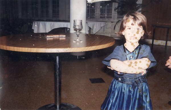 Petite fille brune les bras croisés en robe bleue devant une table.