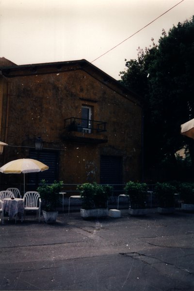 Terrasse de café avec une table, chaises et parasol, plantes en pot, maisons en arrière-plan.