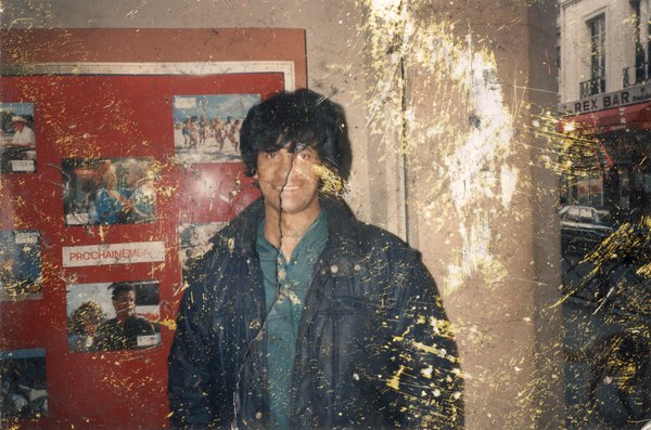 Homme brun debout souriant devant un panneau de photos films à l’entrée d’un cinéma. Image abîmée