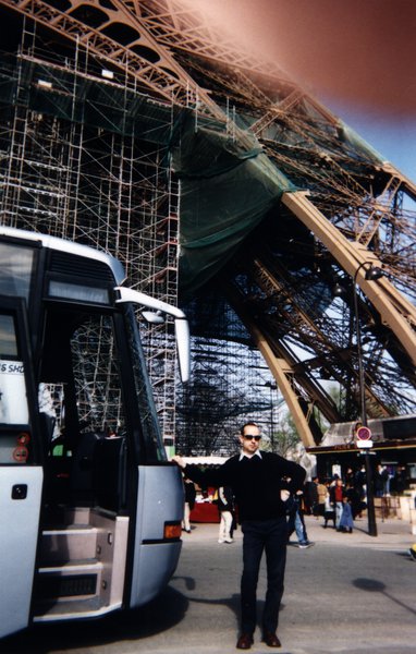 Homme à lunettes noires debout près d’un car touristique sous la Tour Eiffel.