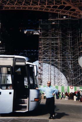 Homme à lunettes noires debout près d’un car touristique sous la Tour Eiffel.