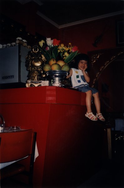Petite fille riant assise de profil sur un comptoir, vase de fleurs et corbeille de fruits en arrière-plan.