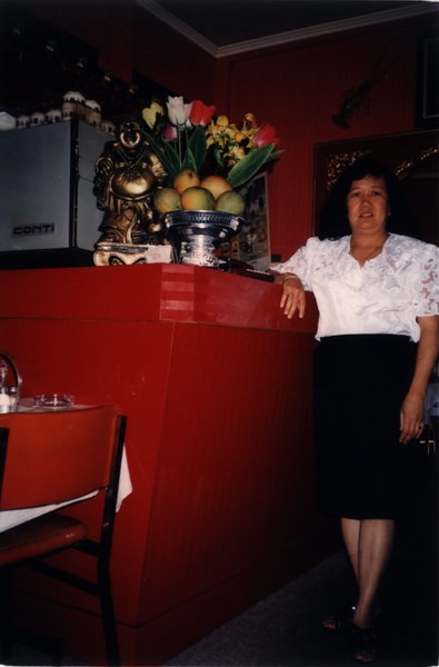 Femme accoudée à un comptoir, corbeille de fruits et vase de fleurs en arrière-plan.