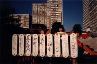 Petite fille debout derrière une palissade blanche, barres d’immeubles en arrière-plan.