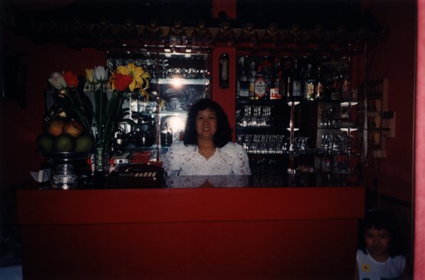 Femme brune souriante en chemisier blanc derrière un comptoir de bar.