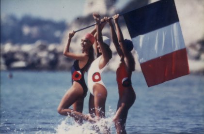 Trois femmes en maillot de bain les pieds dans l’eau avec un drapeau français