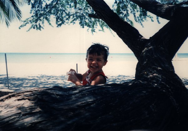 Enfant souriant assis derrière un tronc d’arbre sur une plage.