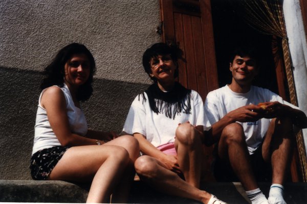 Une femme et deux adolescents assis devant la façade d’une maison.