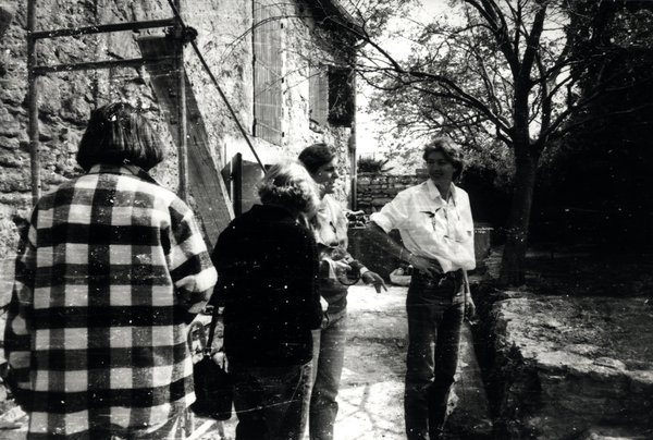 Quatre personnes discutant devant un mur en pierre. Noir et blanc.ᴬᴵ
