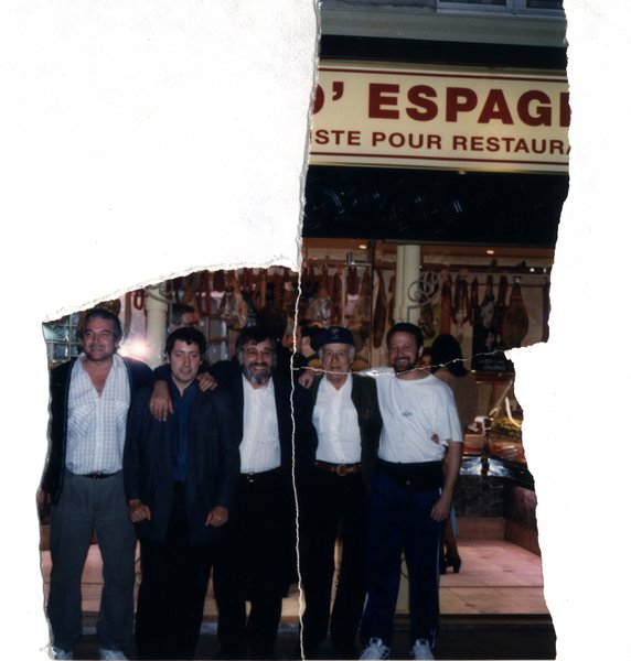 Cinq hommes posant devant une épicerie espagnole avec jambons suspendus. Image avec déchirure, reconstituée.ᴬᴵ