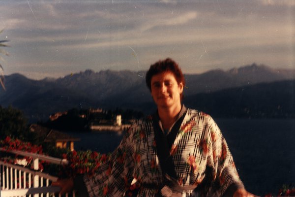 Homme en robe de chambre souriant sur un balcon avec vue sur un lac au coucher du soleil.ᴬᴵ