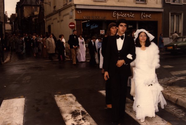 Mariés traversant un passage piéton devant une épicerie fine, invités en cortège.ᴬᴵ