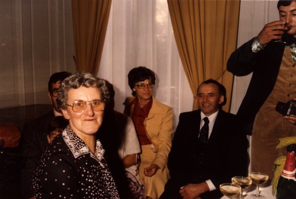 Femme âgée à lunettes au premier plan, groupe assis près d'une fenêtre, coupes de champagne sur la table.ᴬᴵ