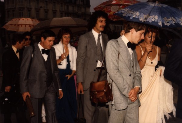 Groupe d'invités en tenue de soirée avec parapluies sur une place.ᴬᴵ