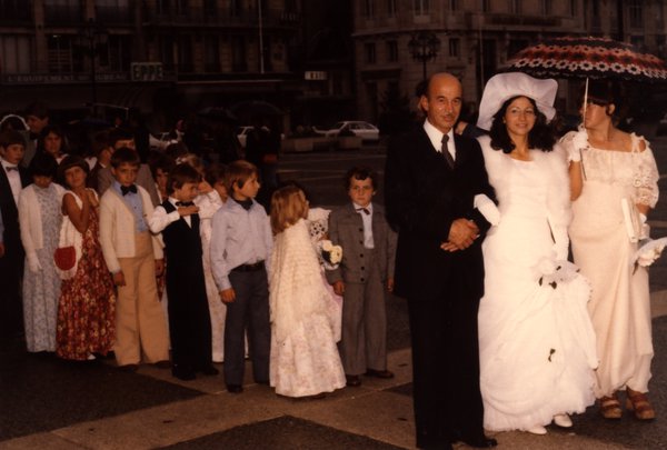 Mariés et enfants d'honneur sur une place, avec parapluie.ᴬᴵ
