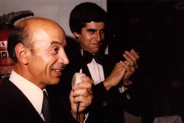 Homme âgé parlant au micro, un autre en nœud papillon derrière lui.ᴬᴵ