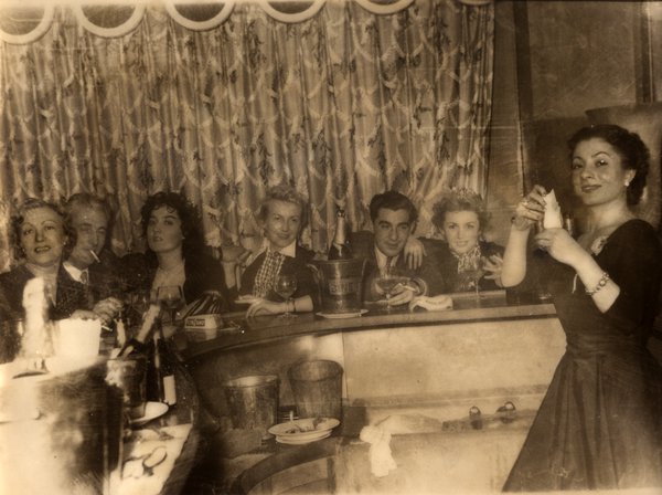Groupe de personnes autour d'un bar, femme debout tenant un objet. Noir et blanc.ᴬᴵ