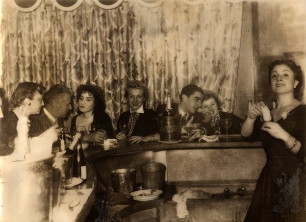 Groupe de personnes autour d'un bar avec seaux à glace. Noir et blanc.ᴬᴵ