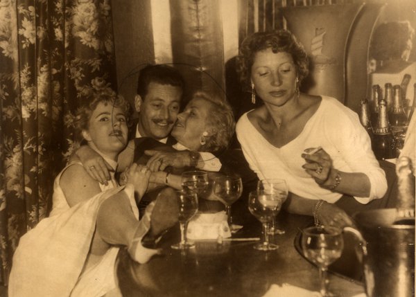 Groupe de quatre personnes enlacées à une table. Noir et blanc.ᴬᴵ