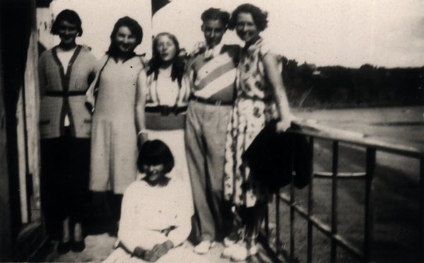 Groupe de jeunes gens sur une terrasse au bord de l'eau, une personne assise au premier plan. Noir et blanc.ᴬᴵ