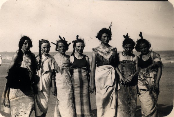 Groupe de femmes en costume avec coiffe à plumes sur une plage. Noir et blanc.ᴬᴵ