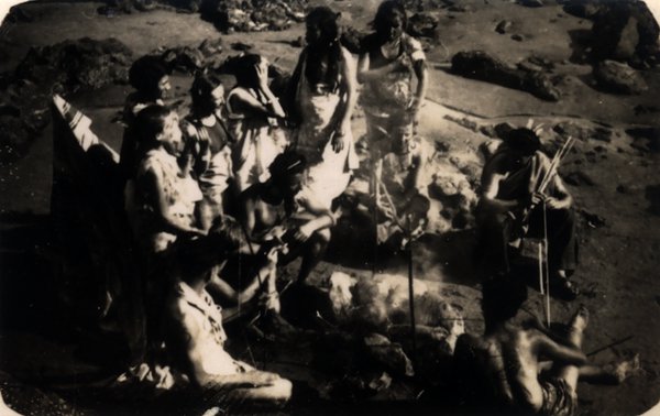 Groupe de personnes déguisées réunies autour d'un feu sur une plage rocheuse. Noir et blanc.ᴬᴵ