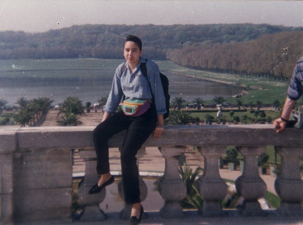 Young brunette girl sitting on a stone balustrade, gardens of the Palace of Versailles in the background.