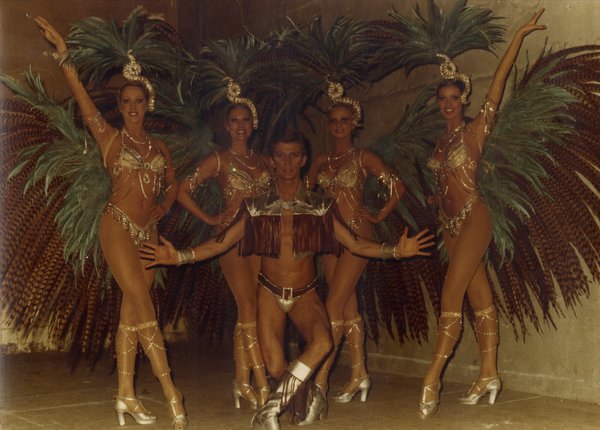 Danseuses de revue avec diadèmes, plumes, et lingeries dorées posent avec un danseur.