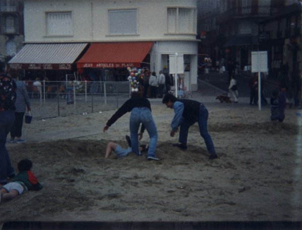 Deux hommes debout et un autre au sol sur le sable, devantures de magasins en arrière-plan.