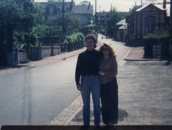 Couple homme brun et femme blonde enlacé dans une rue, maisons en arrière-plan.