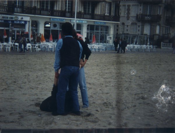 Hommes de dos sur le sable, brasserie et terrasse en arrière-plan.
