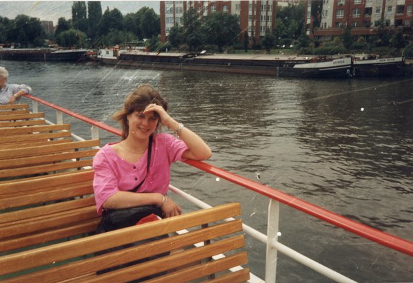 Femme souriante assise sur le pont extérieur d’un bateau-mouche, péniches et immeubles en arrière-plan.