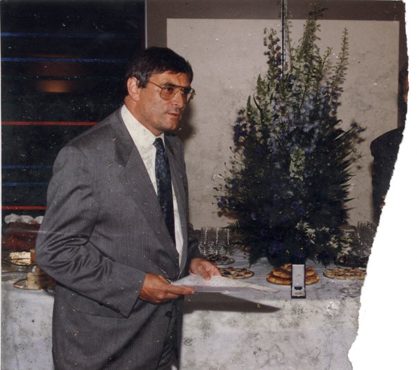 Homme debout en complet gris, une feuille à la main, devant une longue table de buffet. Image déchirée.