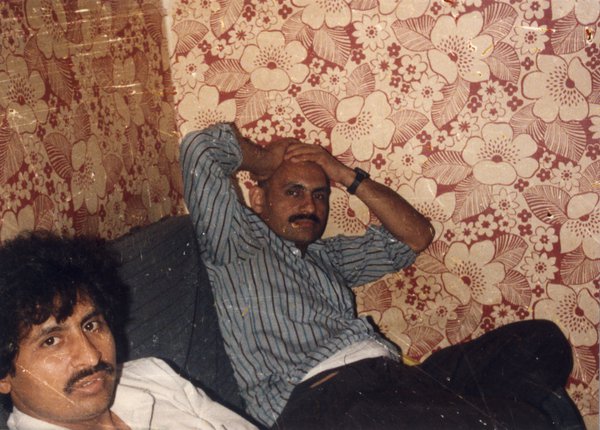 Deux hommes moustachus assis sur un canapé, mur avec papier peint en arrière-plan.