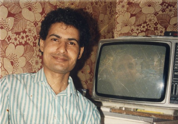 Visage d’homme souriant devant un téléviseur, mur recouvert de papier peint en arrière-plan.