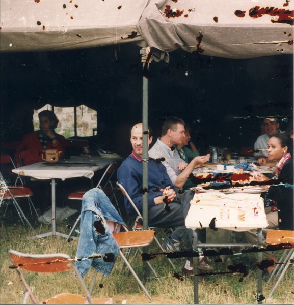 Hommes et femmes autour d’un repas posé sur des tables pliantes sous une tente.  Images avec traces de rayure.
