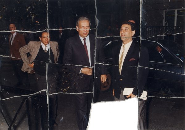 Trois hommes en complet debout devant une voiture noire. Image déchirée, en partie reconstituée.