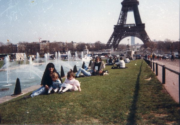 Femme et deux enfants assis sur l'herbe devant la tour Eiffel et des fontaines.ᴬᴵ