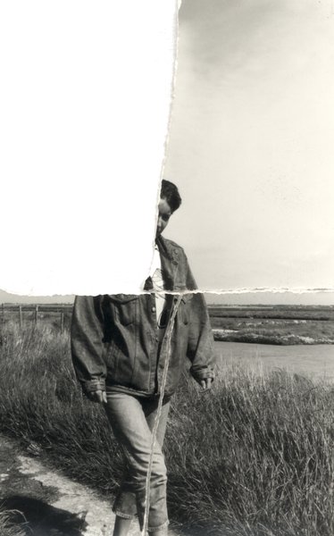 Femme debout dans un chemin de campagne avec un blouson en jean, plan d’eau en arrière-plan. Noir et blanc. Image déchirée en partie reconstituée.