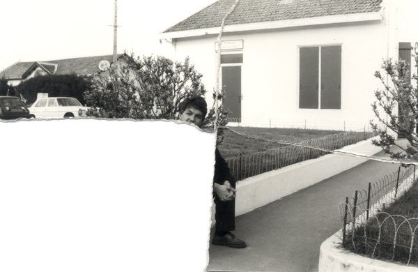 Homme assis devant un pavillon. Noir et blanc. Image déchirée en partie reconstituée.