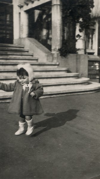 Enfant en manteau et bonnet devant le perron d’une maison. Noir et blanc.
