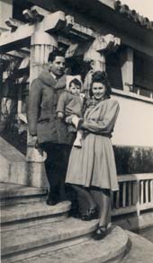 Couple avec un enfant dans les bras pose en haut du perron d’une maison. Noir et blanc.